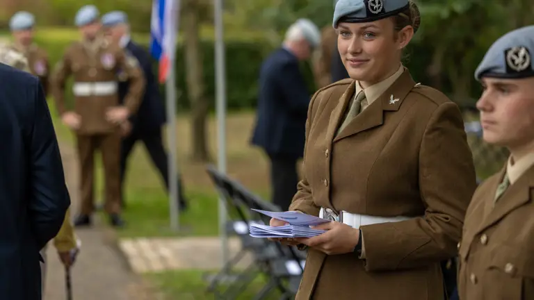 A person in a brown military dress uniform with a light blue beret holds a stack of papers, standing outdoors at a formal event. Other uniformed individuals and a flag are visible in the background.