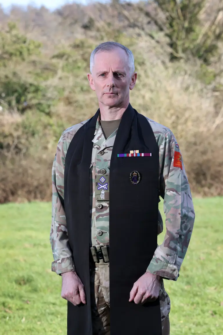 A man in military uniform stands outdoors on grass, wearing a long black scarf with badges. The background is blurred trees under clear skies. He has a serious expression.