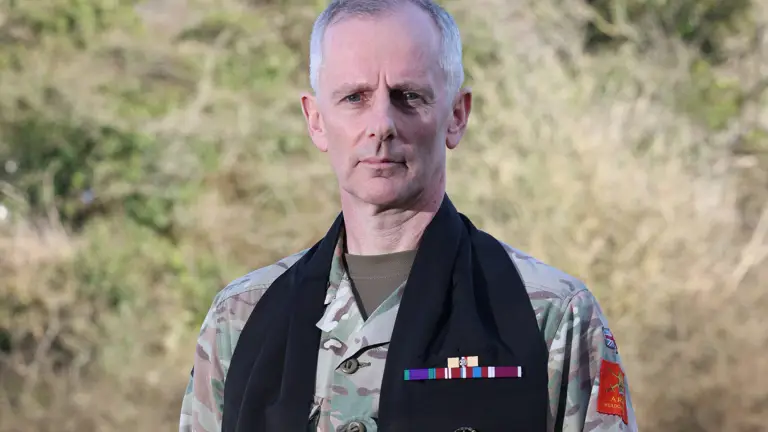 A man in military uniform stands outdoors on grass, wearing a long black scarf with badges. The background is blurred trees under clear skies. He has a serious expression.