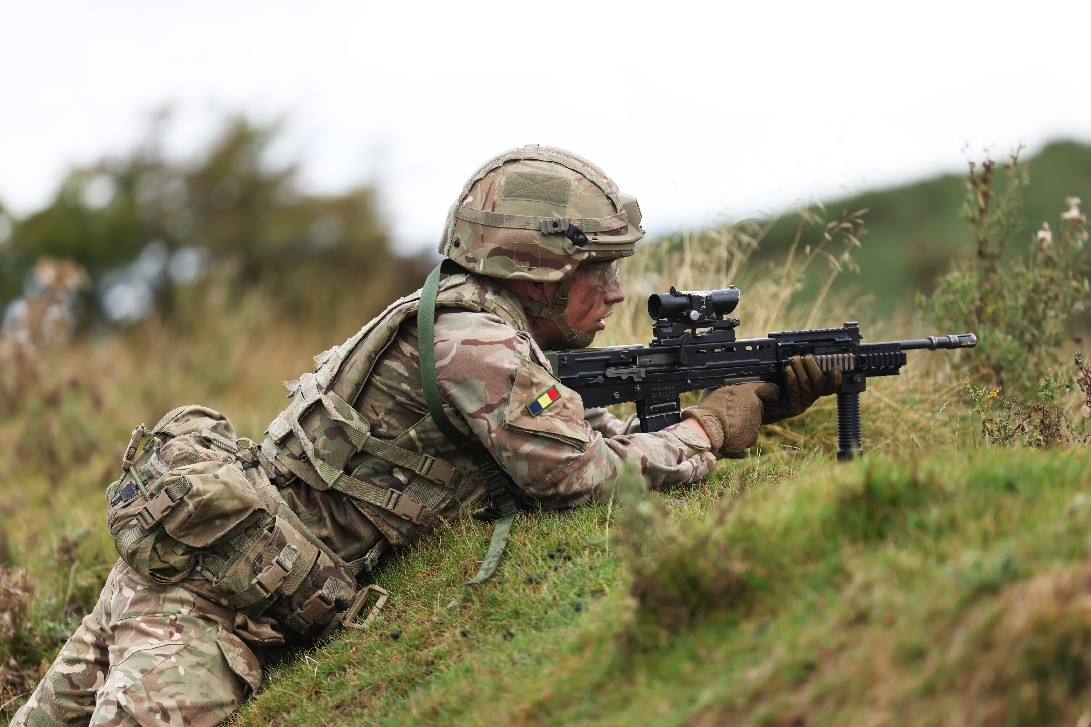 Soldier in camouflage uniform lying prone on grassy hill aiming a scoped rifle during training exercise.