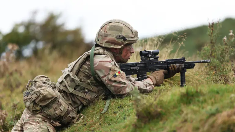 Soldier in camouflage uniform lying prone on grassy hill aiming a scoped rifle during training exercise.