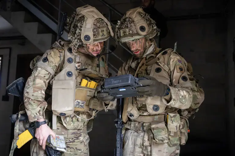 Two soldiers in full tactical gear operate a handheld drone controller inside a dimly lit industrial building.