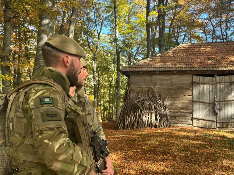Two soldiers in camouflage uniforms look on at a building.