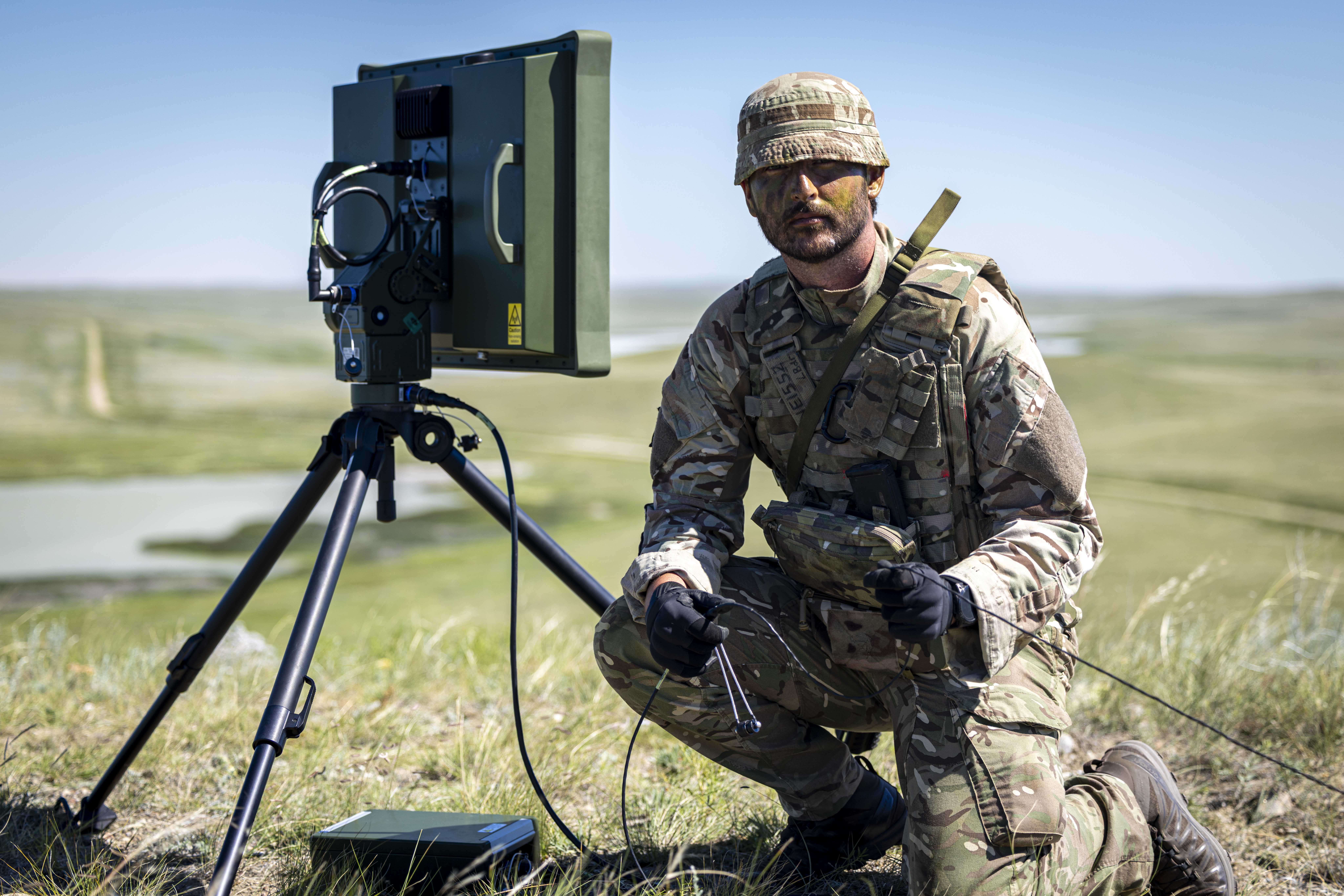 A person in camouflage military attire is kneeling beside a portable radar system on a tripod in an open grassy field. The landscape features distant rolling hills and a body of water. The person is holding a small device connected to the radar system.