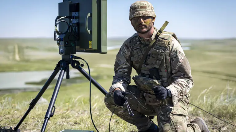 A person in camouflage military attire is kneeling beside a portable radar system on a tripod in an open grassy field. The landscape features distant rolling hills and a body of water. The person is holding a small device connected to the radar system.