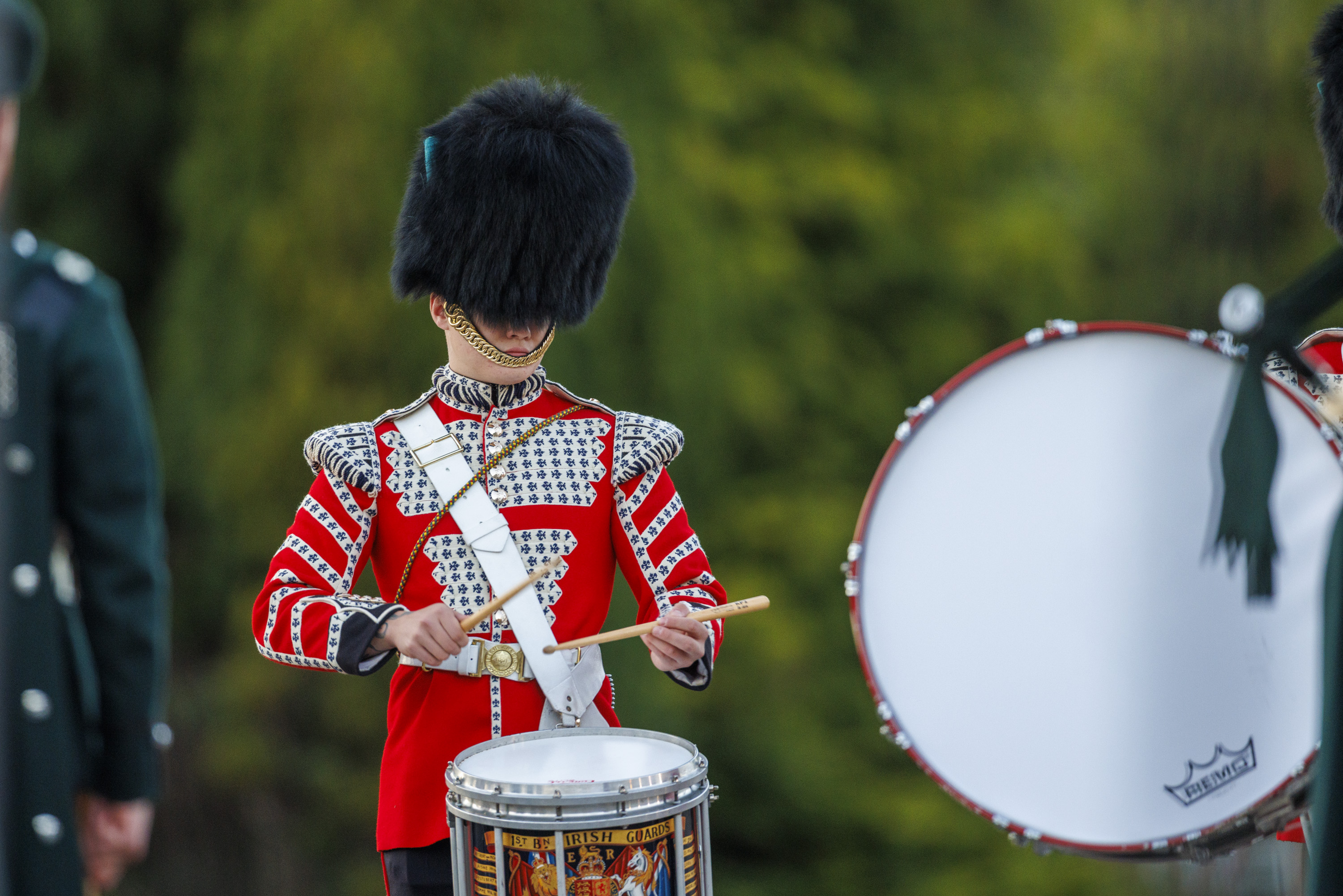 A drummer in a red and white ceremonial uniform playing a drum with a detailed crest on it outdoors.