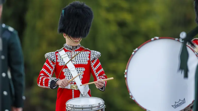 A drummer in a red and white ceremonial uniform playing a drum with a detailed crest on it outdoors.