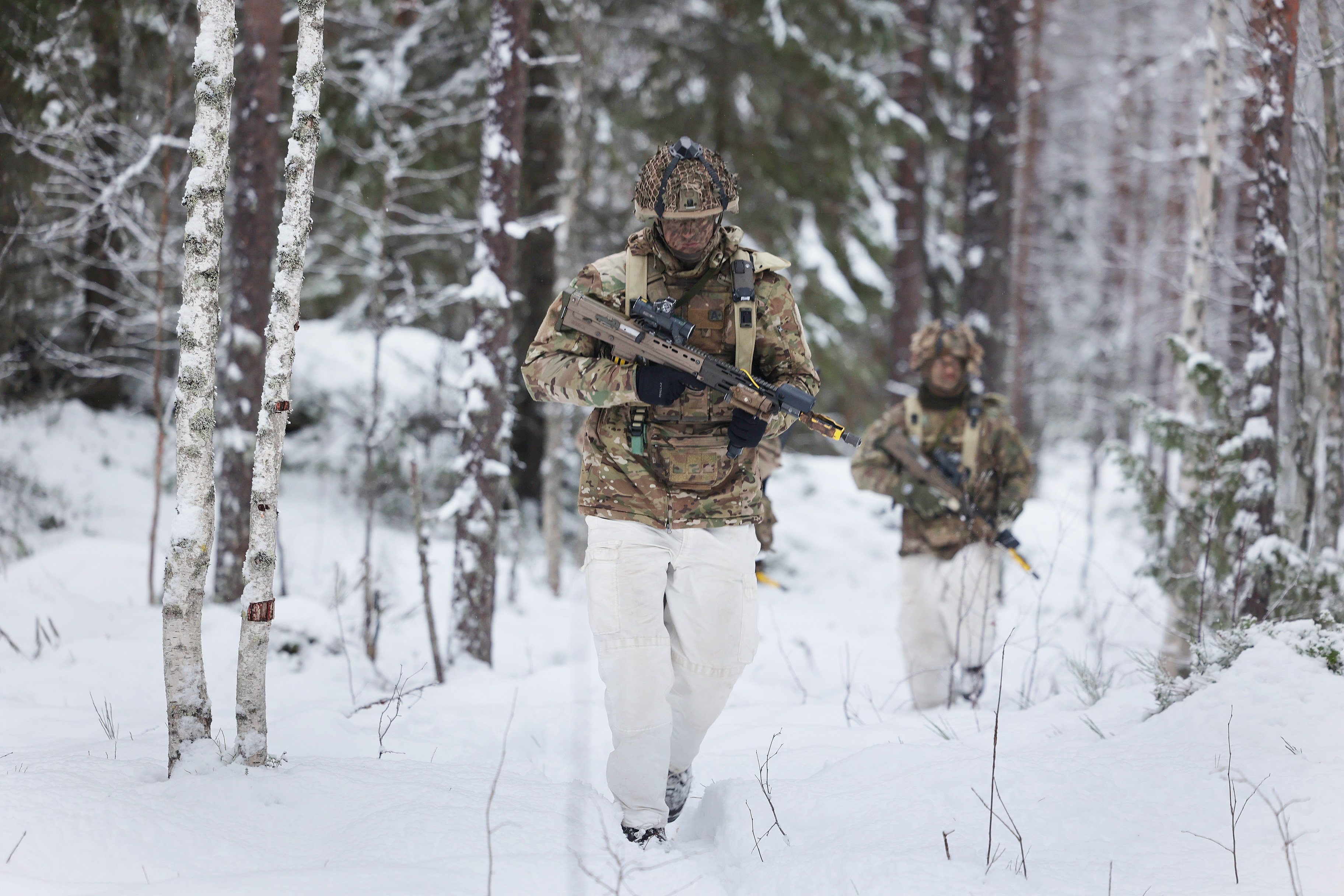 Two soldiers in winter camouflage walk through a snowy forest alongside camouflaged military vehicles, creating a scene of focused, cold readiness.