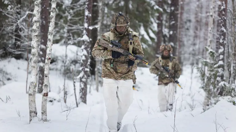 Two soldiers in winter camouflage walk through a snowy forest alongside camouflaged military vehicles, creating a scene of focused, cold readiness.