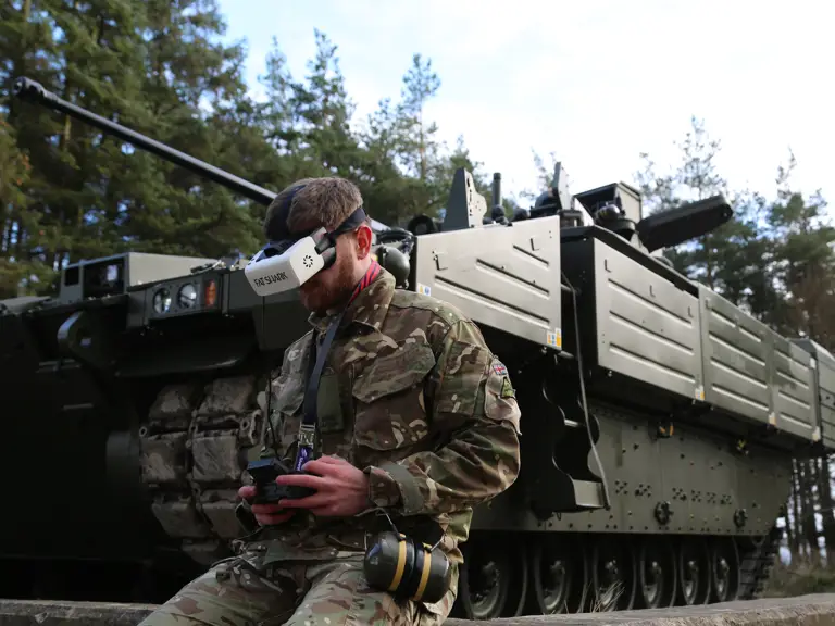 A soldier from the Royal Lancers wears a virtual reality headset while kneeling in front of a green armoured vehicle.