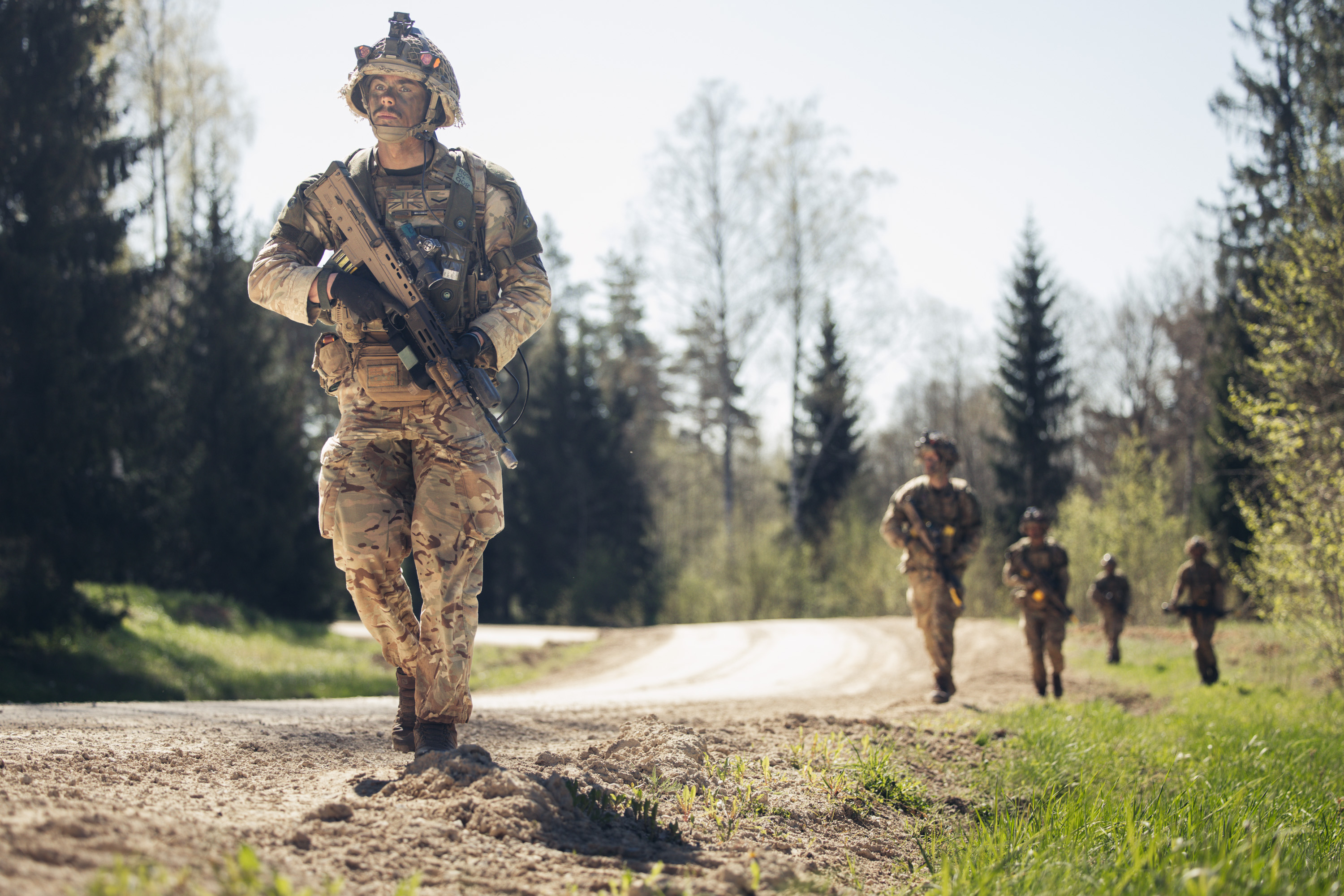 Several soldiers in uniform walk with their weapons against a woodland backdrop.