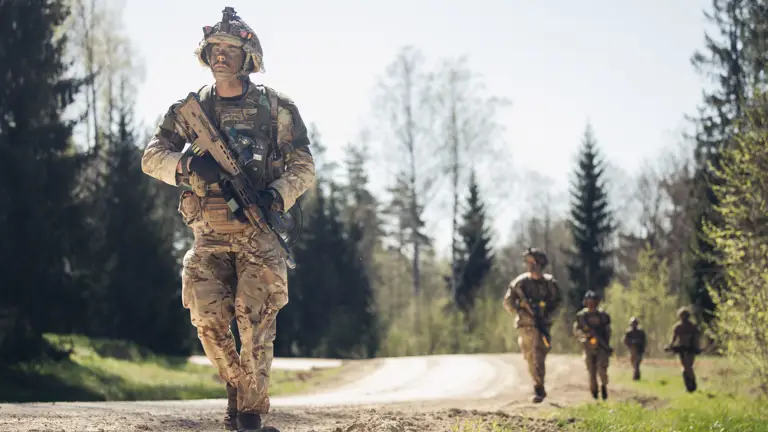 Several soldiers in uniform walk with their weapons against a woodland backdrop.