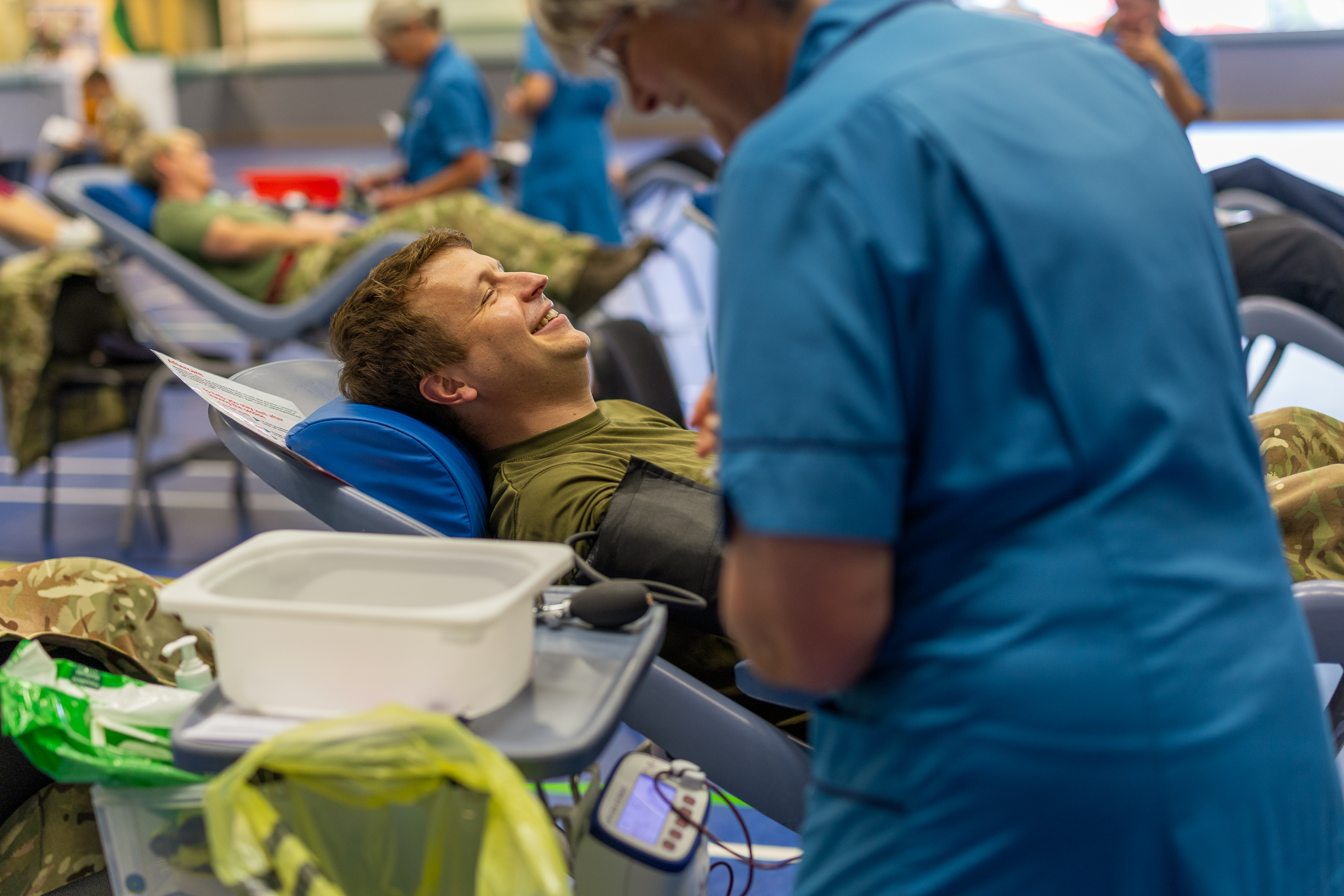 Military personnel lie on reclining chairs donating blood in a clinical setting with medical staff attending to them.