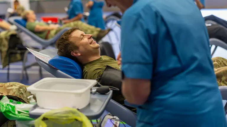 Military personnel lie on reclining chairs donating blood in a clinical setting with medical staff attending to them.