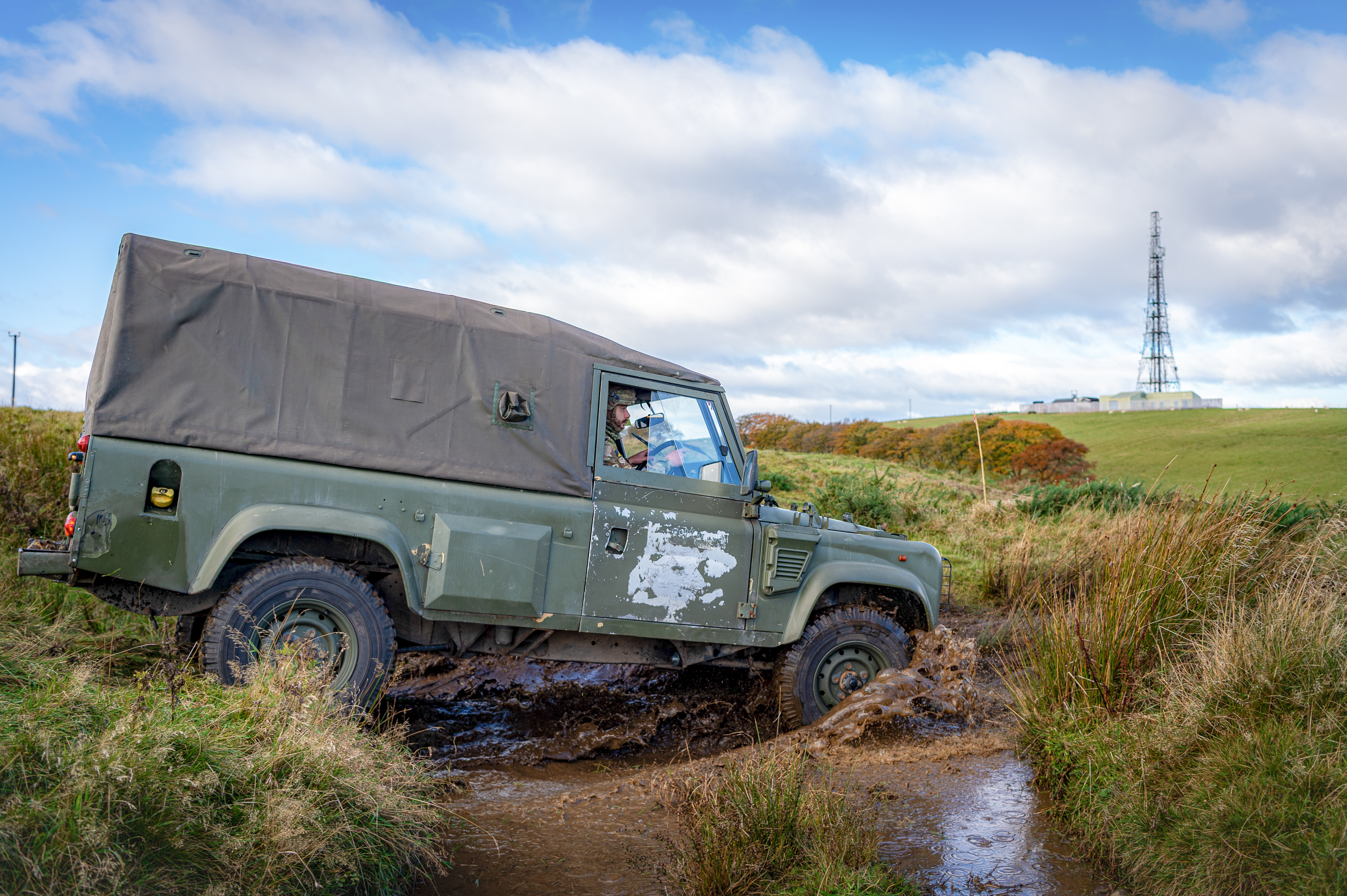 A rugged green off-road vehicle traverses a muddy path in a grassy field. A radio tower stands on a hill under a cloudy sky in the background.