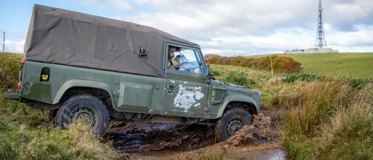 A rugged green off-road vehicle traverses a muddy path in a grassy field. A radio tower stands on a hill under a cloudy sky in the background.