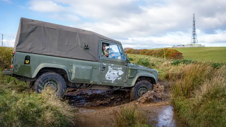 A rugged green off-road vehicle traverses a muddy path in a grassy field. A radio tower stands on a hill under a cloudy sky in the background.