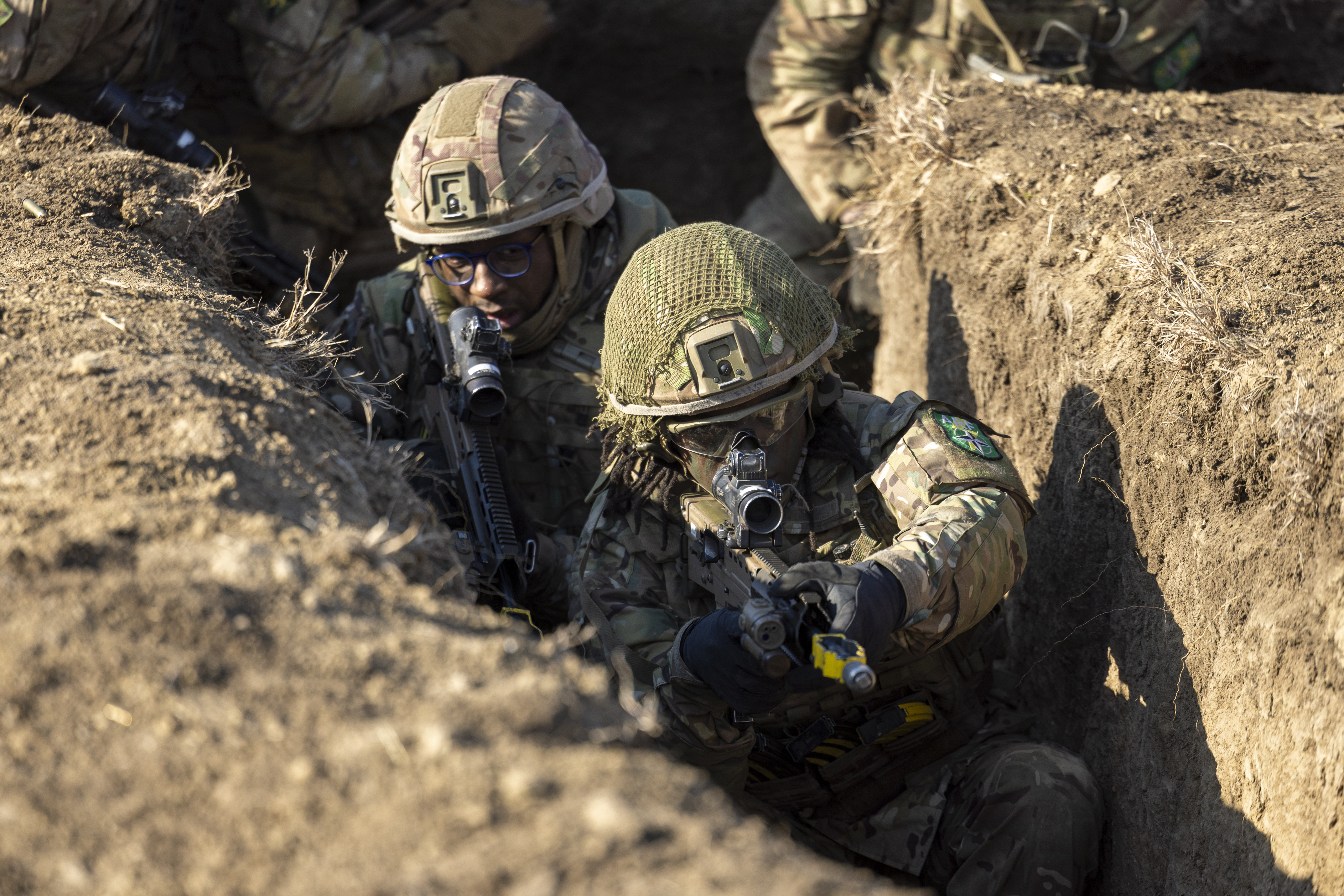 Two soldiers in camouflage uniform seen clearing a trench during exercise with their rifles at the ready. 