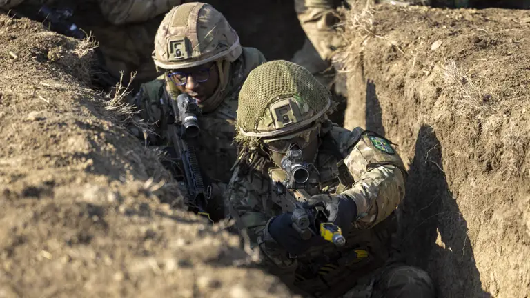 Two soldiers in camouflage uniform seen clearing a trench during exercise with their rifles at the ready.