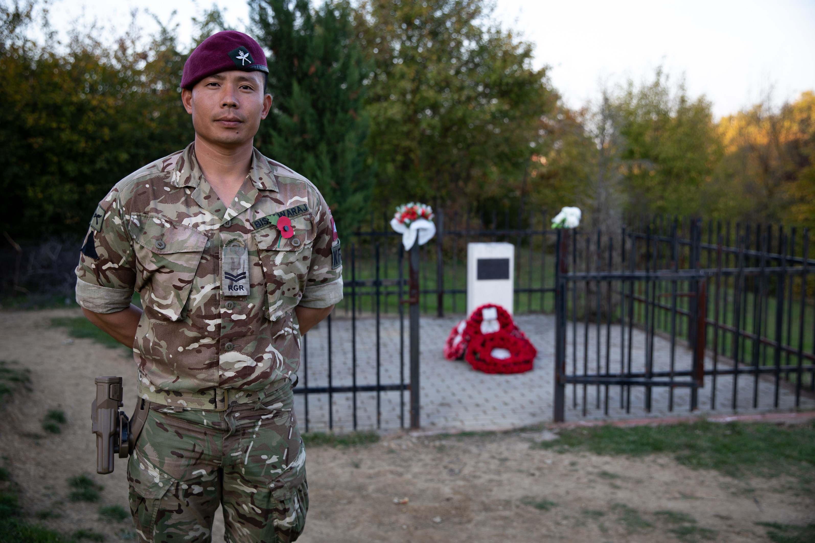 A soldier wearing camouflage uniform and a maroon beret stands in front of a memorial.