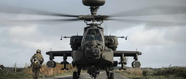 Front view of a military Apache helicopter with spinning rotor blades on a rural road beside a soldier in camouflage gear.
