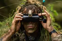 A person with face paint and vegetation in their clothing is using a pair of FPV goggles outdoors.