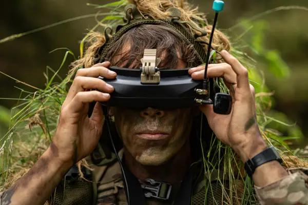 A person with face paint and vegetation in their clothing is using a pair of FPV goggles outdoors.