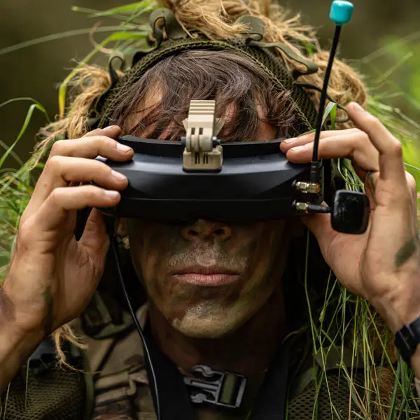 A person with face paint and vegetation in their clothing is using a pair of FPV goggles outdoors.