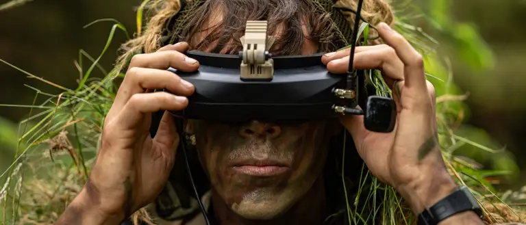 A person with face paint and vegetation in their clothing is using a pair of FPV goggles outdoors.