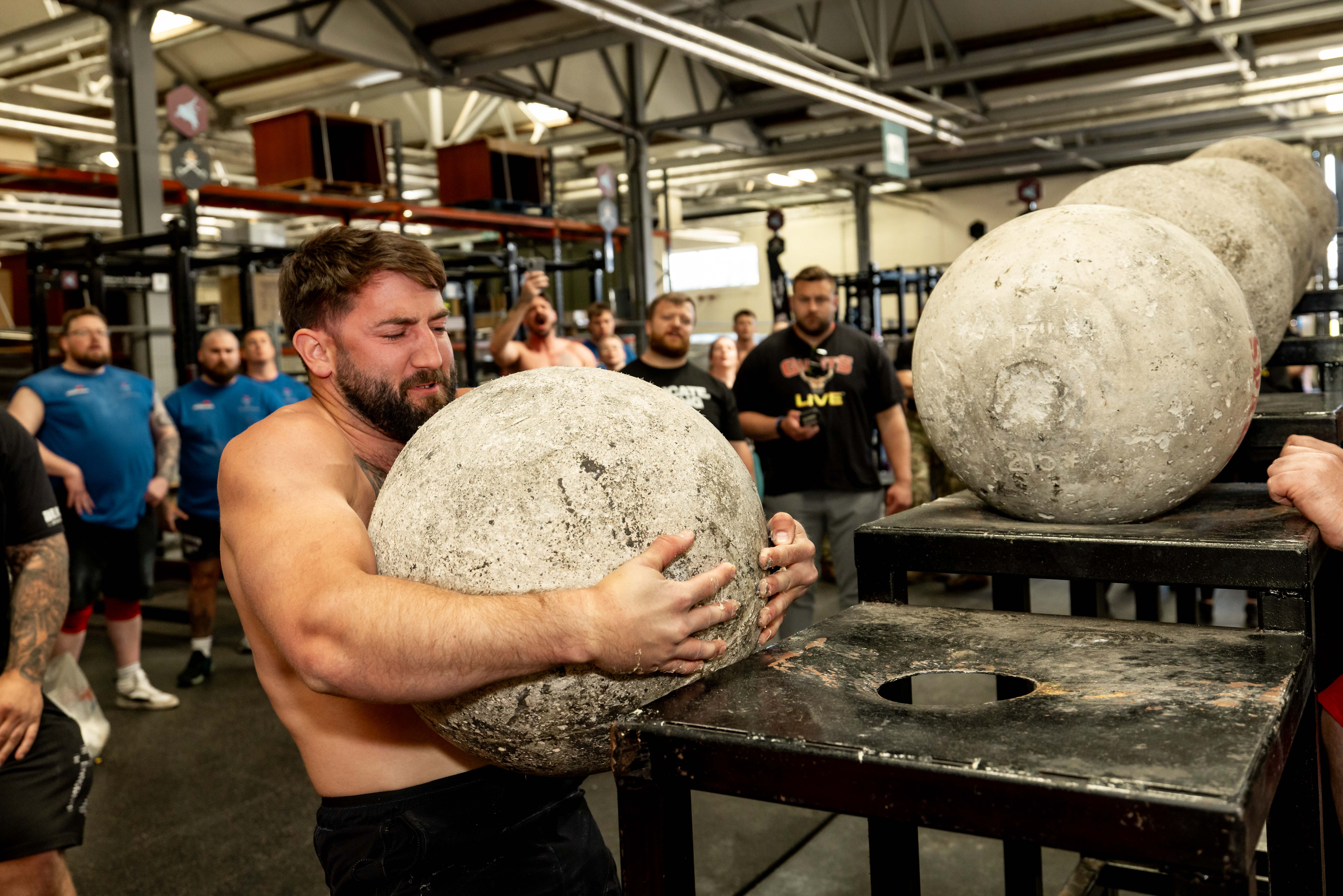 A man strains to lift a large concrete atlas stone onto a platform in a gym. Onlookers watch attentively, adding a sense of tension and focus.