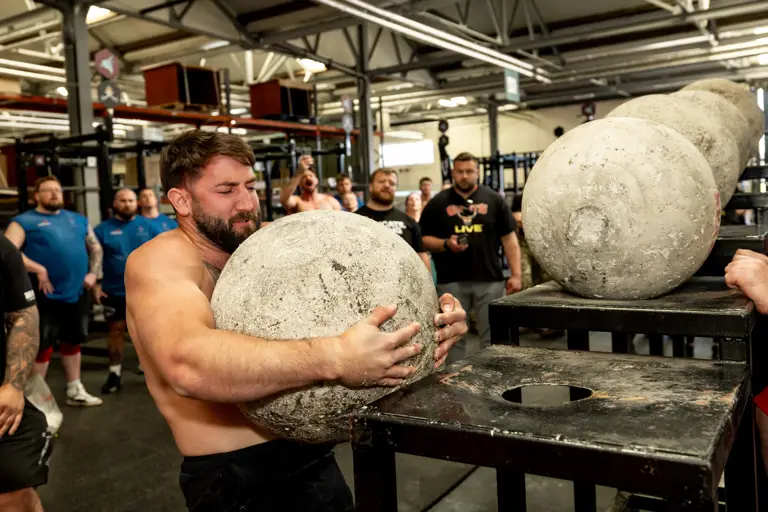 A man strains to lift a large concrete atlas stone onto a platform in a gym. Onlookers watch attentively, adding a sense of tension and focus.