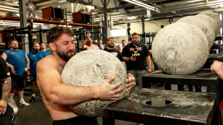 A man strains to lift a large concrete atlas stone onto a platform in a gym. Onlookers watch attentively, adding a sense of tension and focus.