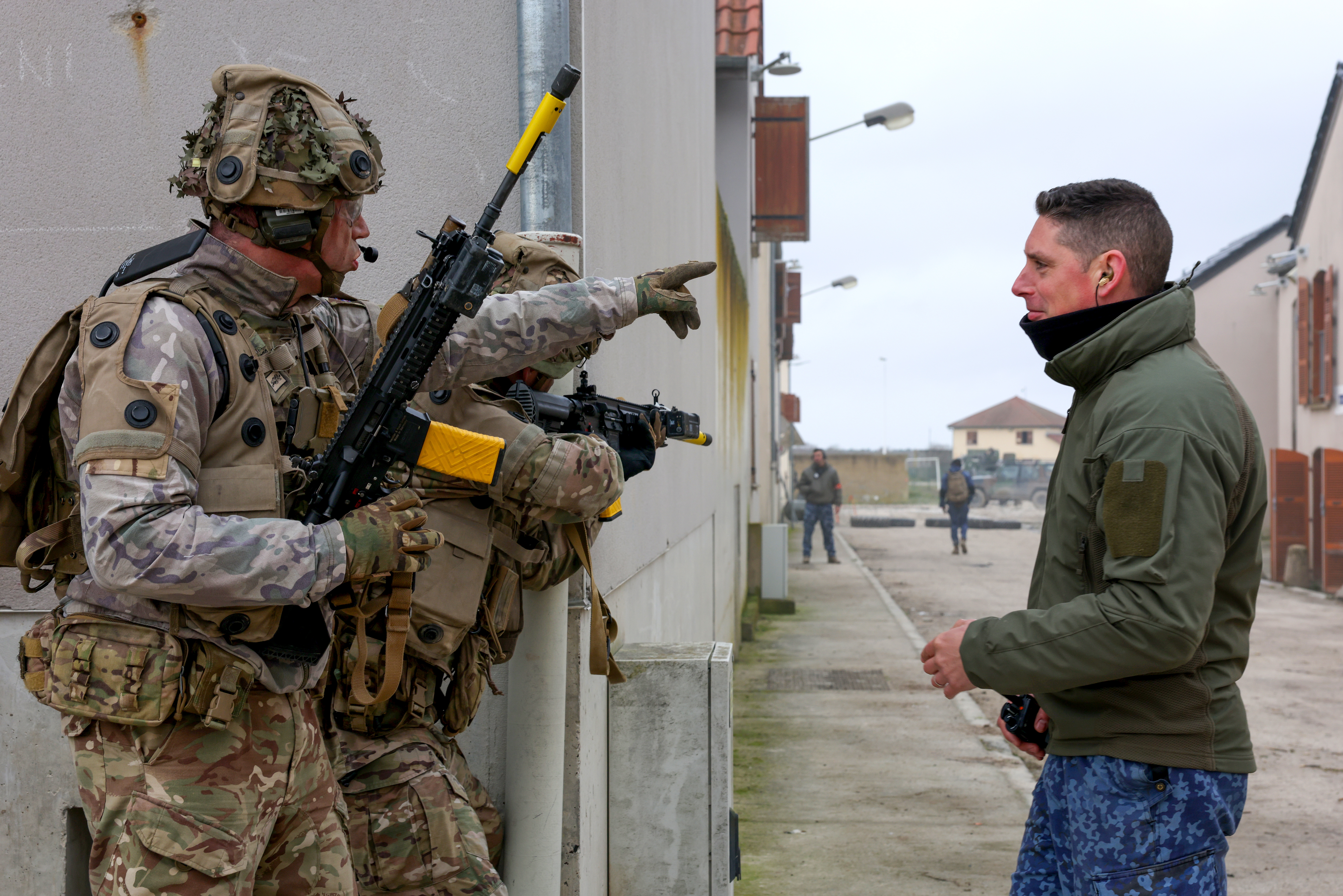 Two soldiers in camouflage gear talking to a man in a green jacket standing on a narrow street between buildings.