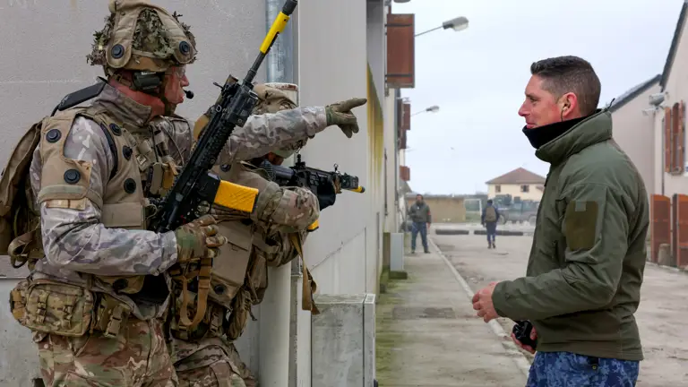 Two soldiers in camouflage gear talking to a man in a green jacket standing on a narrow street between buildings.