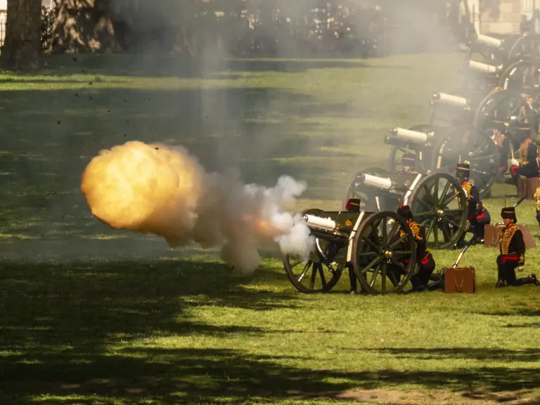 A ceremonial volley of cannons firing on a grassy field, with smoke and fire emerging from one of the cannons.
