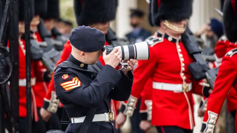 A staff sergeant army photographer dressed in his Number 1 uniform takes photos of a parade of guardsman as they march past him. The guardsman are wearing their red tunics and bearskins.