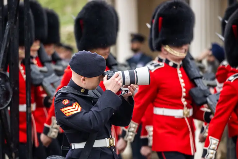 A staff sergeant army photographer dressed in his Number 1 uniform takes photos of a parade of guardsman as they march past him. The guardsman are wearing their red tunics and bearskins.