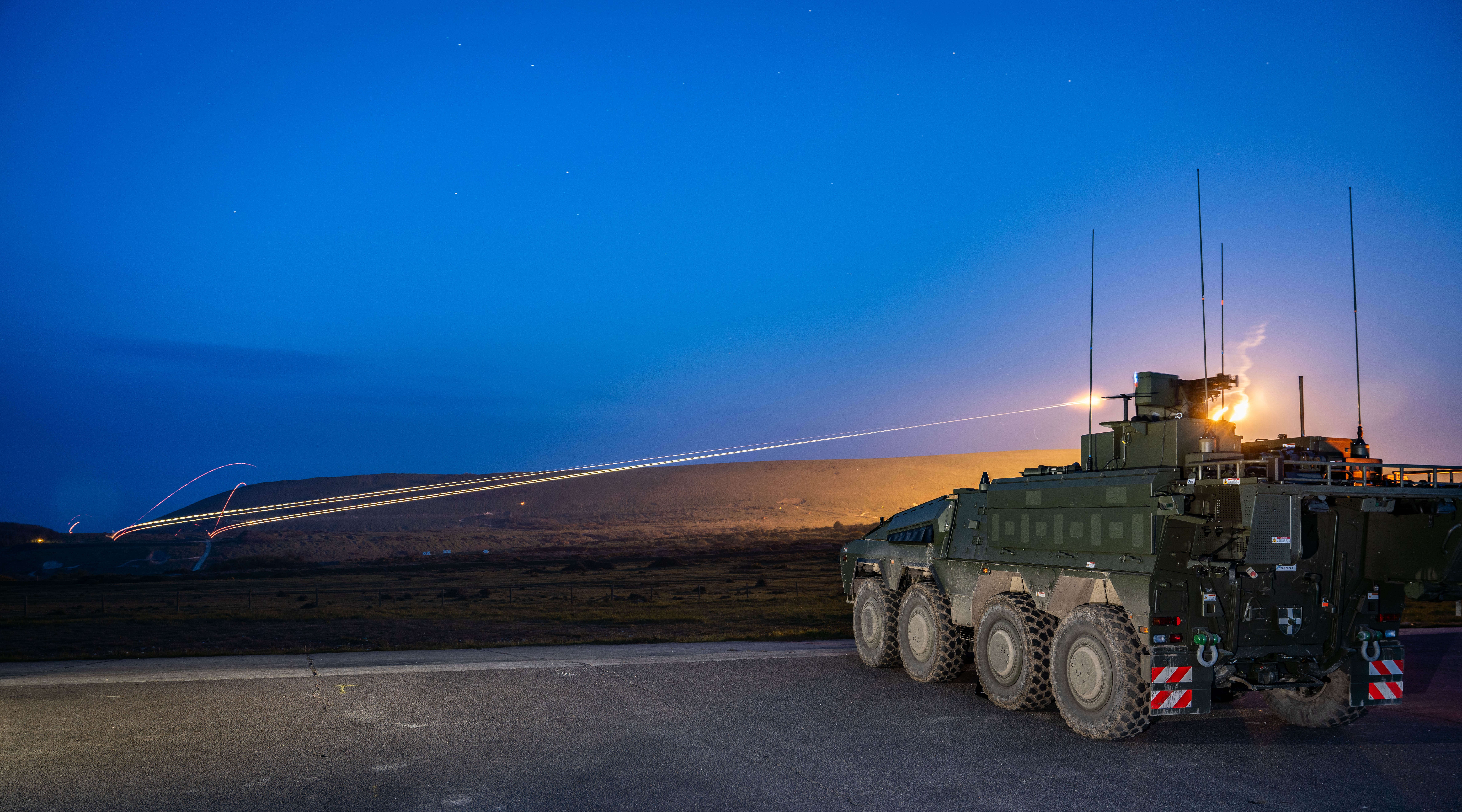 A military vehicle is stationed on a road at dusk. The sky is a deep blue, and distant hills are visible.