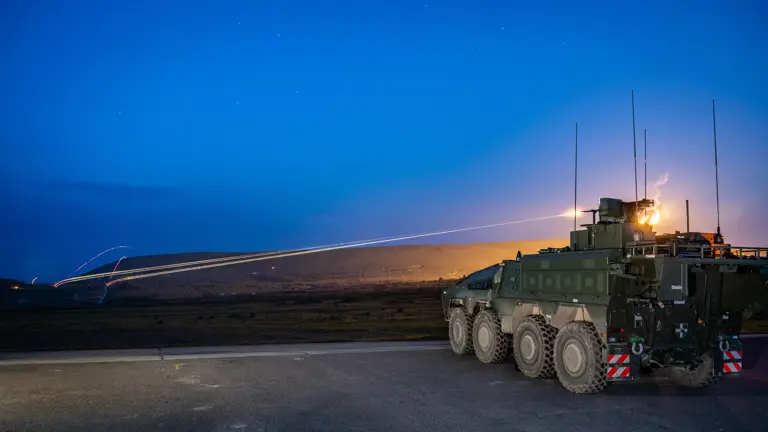 A military vehicle is stationed on a road at dusk. The sky is a deep blue, and distant hills are visible.