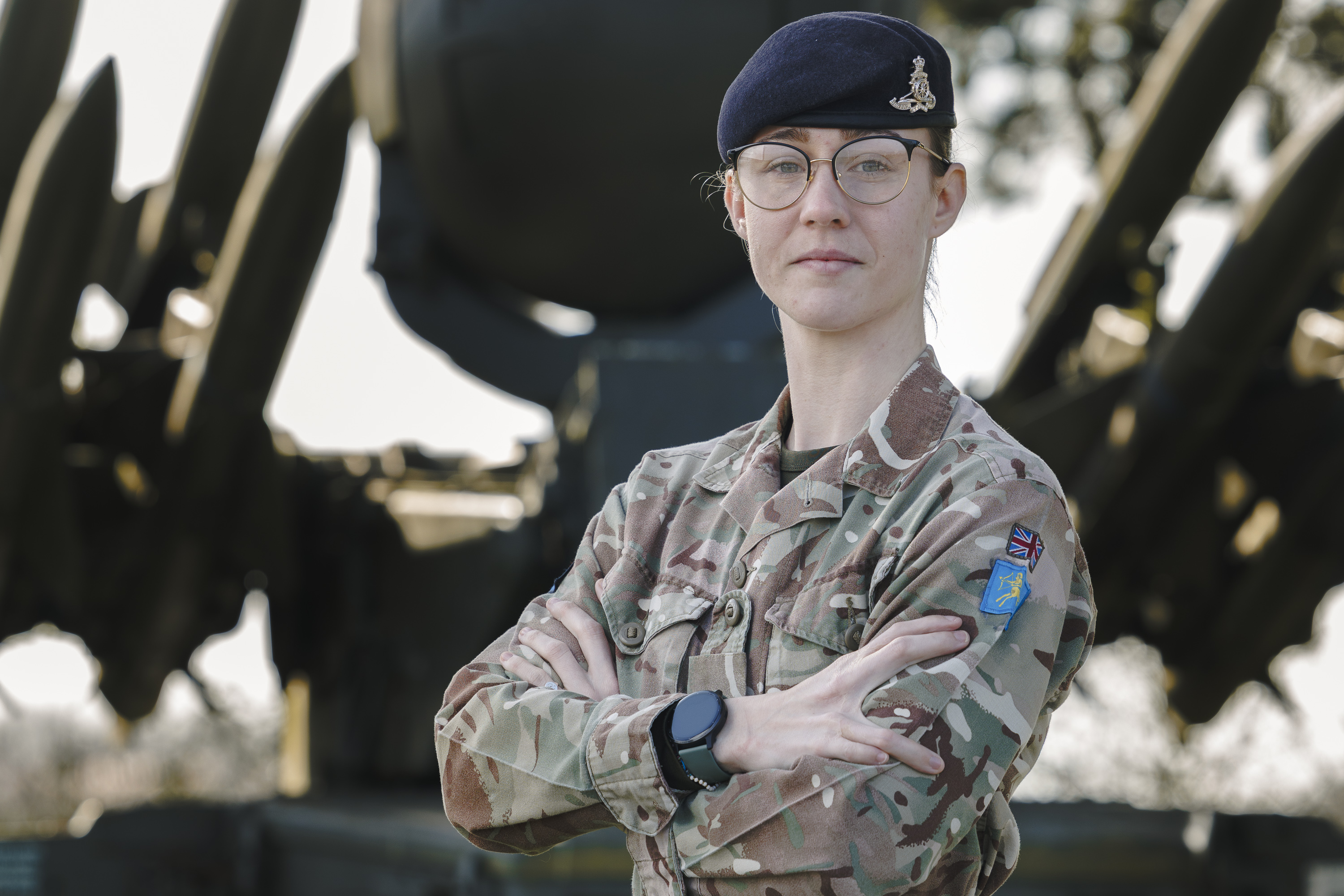 An individual in military camouflage stands confidently with arms crossed in front of a military vehicle equipped with missiles. The background includes grass and trees.