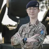 An individual in military camouflage stands confidently with arms crossed in front of a military vehicle equipped with missiles. The background includes grass and trees.