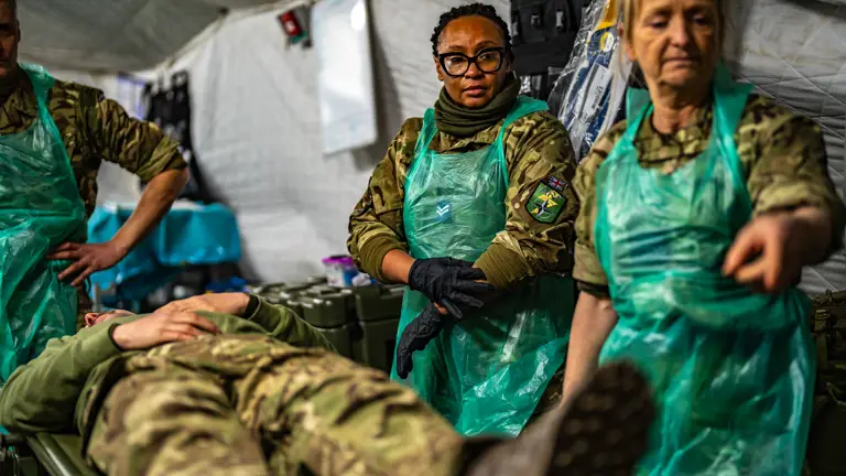 Three Medics in camouflage uniform and PPE are seen treating a wounded Soldier laying down on a stretcher also in camouflage uniform in a field hospital.