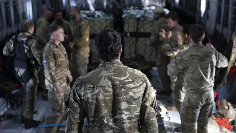 Soldiers in camouflage uniforms stand inside a military cargo plane, engaging in conversation. The atmosphere is focused and collaborative. Parachutes are visible.