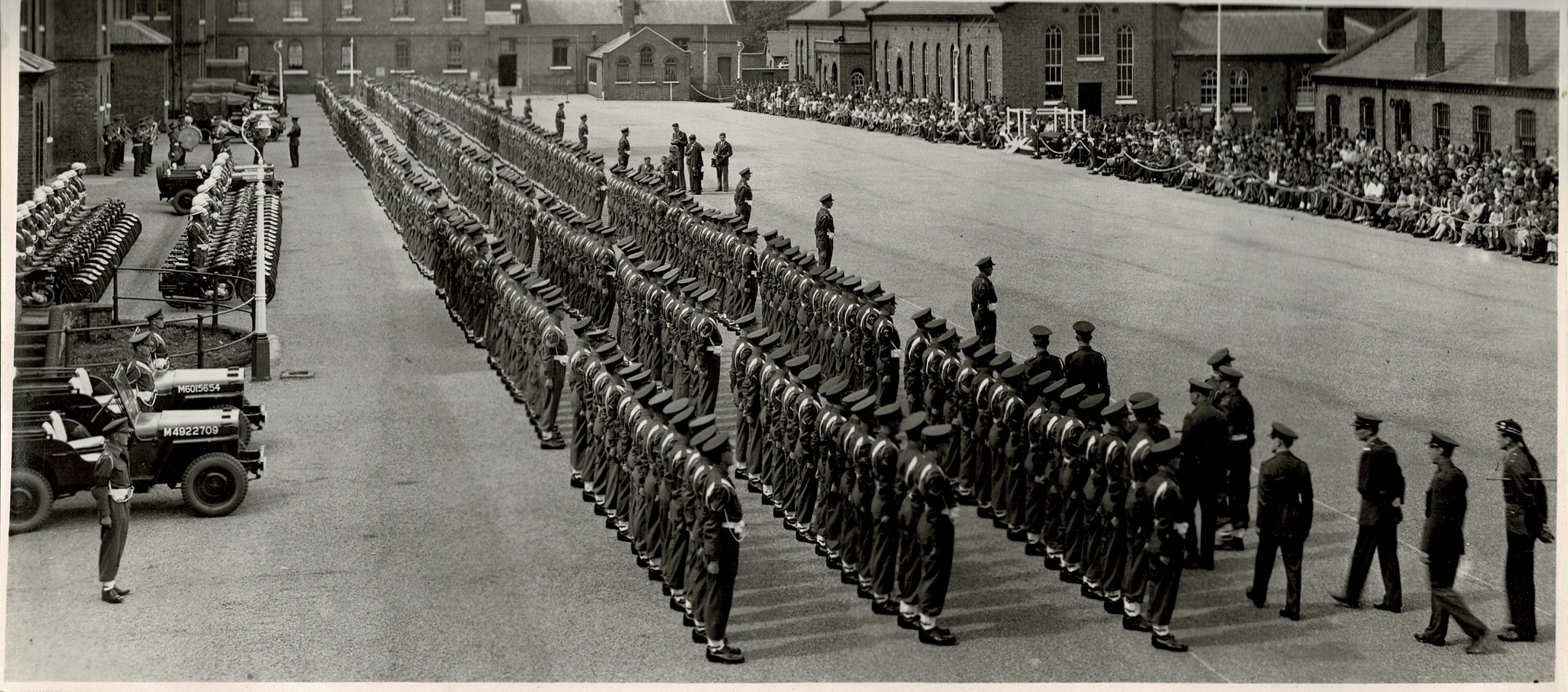 A large formation of military personnel stands in organized rows on a parade ground. Several military vehicles, including jeeps, are lined up on the left side. Buildings surround the area, with spectators seated along the edges. The scene conveys a sense of formality and organisation.