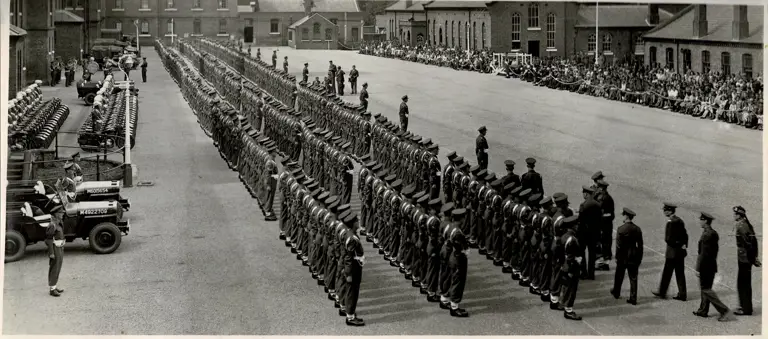 A large formation of military personnel stands in organized rows on a parade ground. Several military vehicles, including jeeps, are lined up on the left side. Buildings surround the area, with spectators seated along the edges. The scene conveys a sense of formality and organisation.