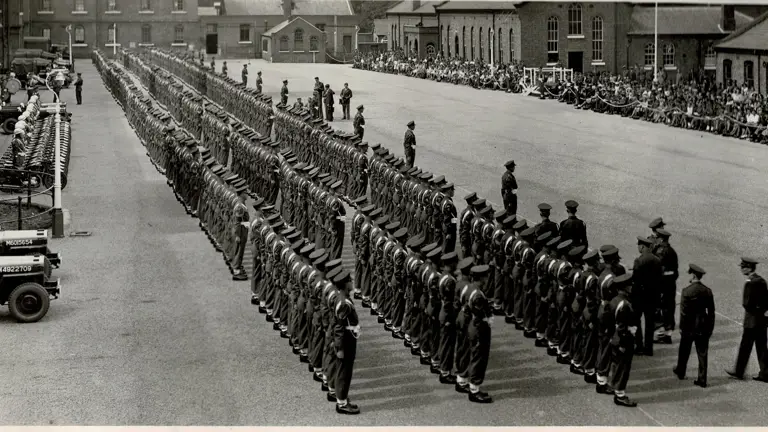 A large formation of military personnel stands in organized rows on a parade ground. Several military vehicles, including jeeps, are lined up on the left side. Buildings surround the area, with spectators seated along the edges. The scene conveys a sense of formality and organisation.