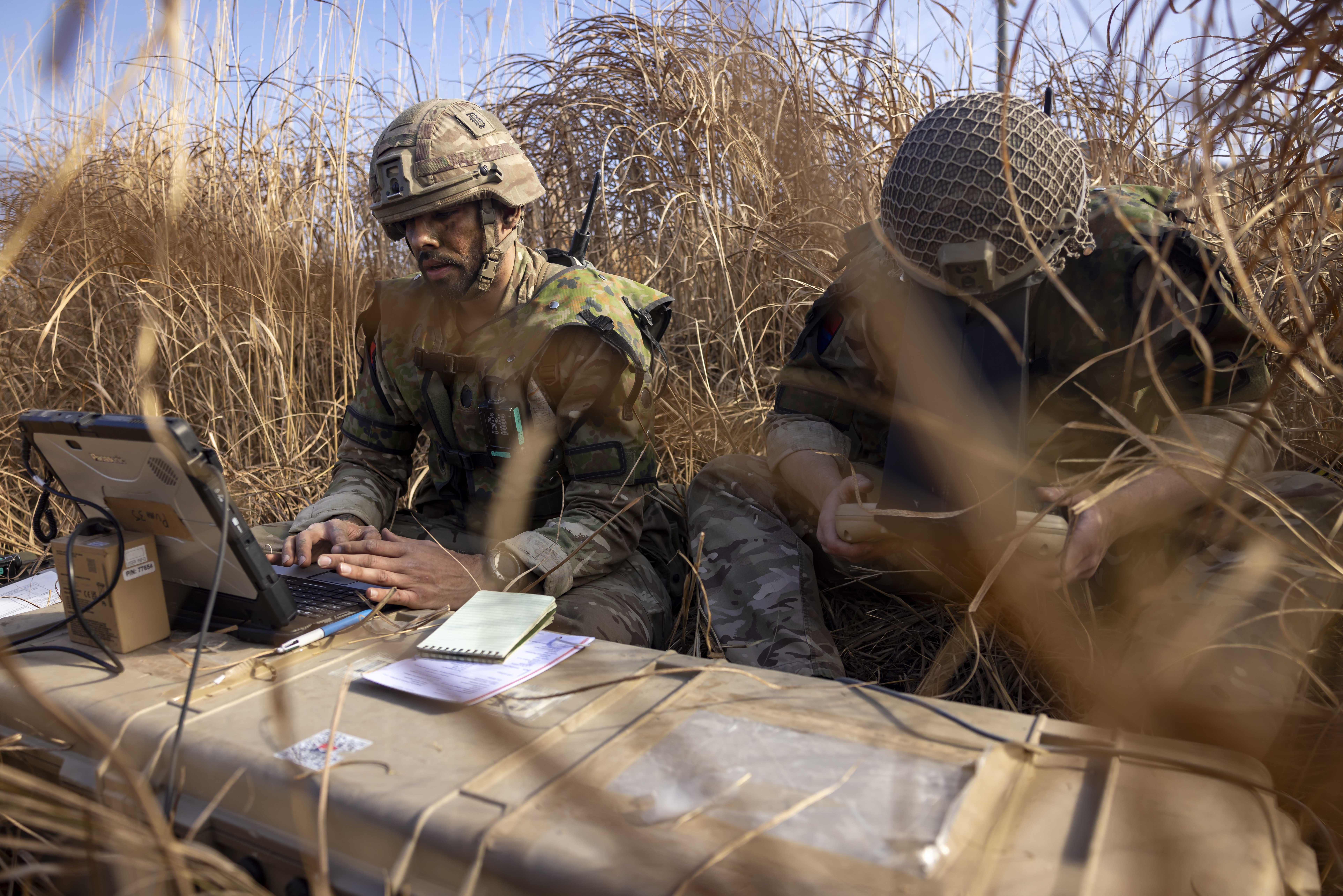 Two British soldiers in camouflage unifoom sat in a grassy terrain on exercise are seen to guide the unmanned aircraft systems “Puma” with specialised equipment. 