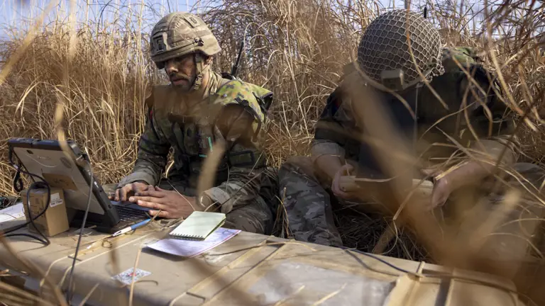 Two British soldiers in camouflage unifoom sat in a grassy terrain on exercise are seen to guide the unmanned aircraft systems “Puma” with specialised equipment.