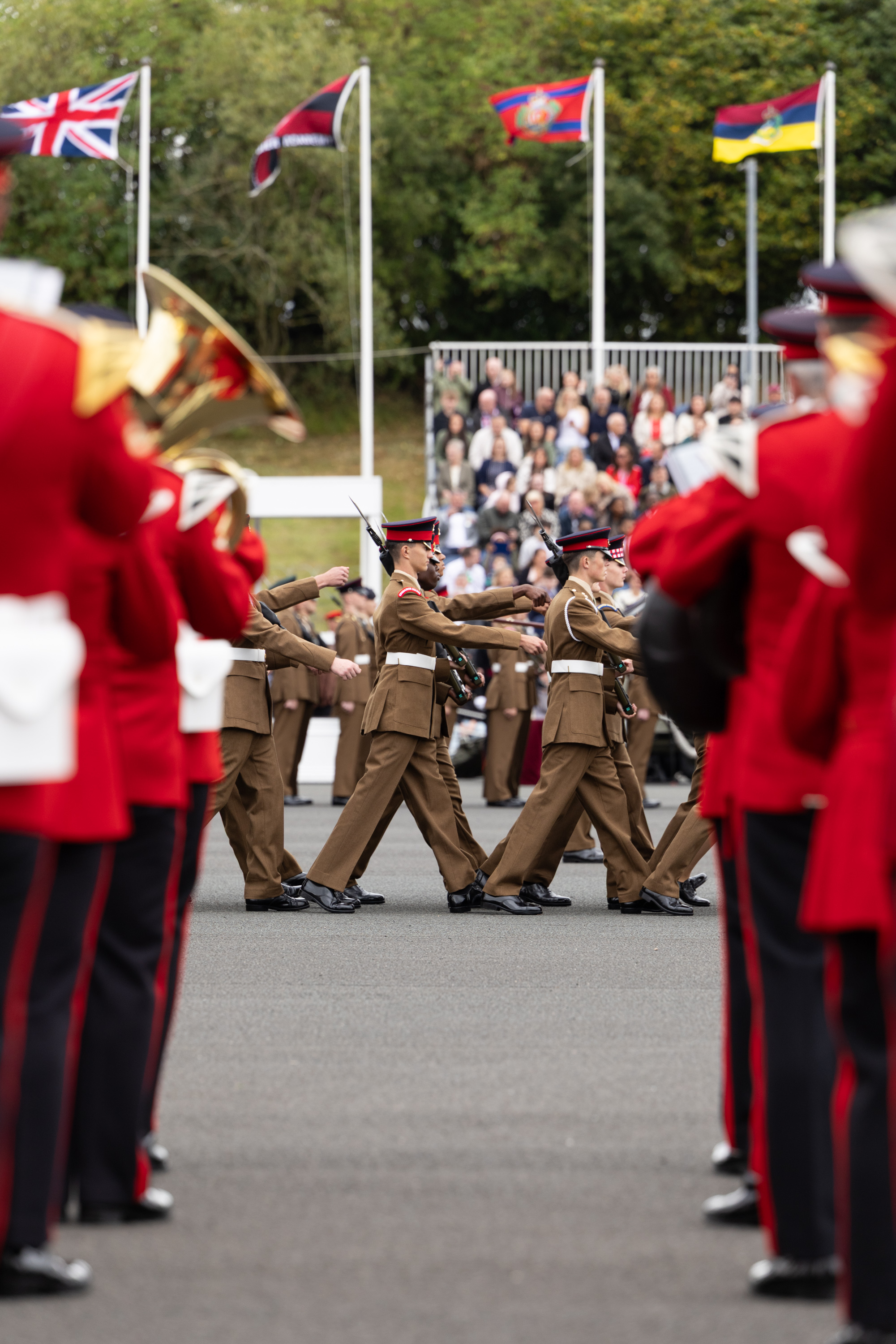 A group of uniformed individuals marches in formation during a parade. They are surrounded by other uniformed individuals playing instruments, dressed in red uniforms. Flags, including the Union Jack, are displayed in the background. Spectators are seated on raised stands observing the event.