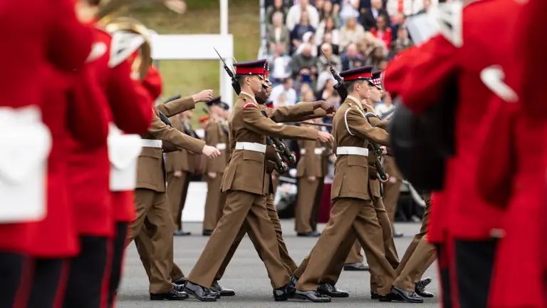 A group of uniformed individuals marches in formation during a parade. They are surrounded by other uniformed individuals playing instruments, dressed in red uniforms. Flags, including the Union Jack, are displayed in the background. Spectators are seated on raised stands observing the event.